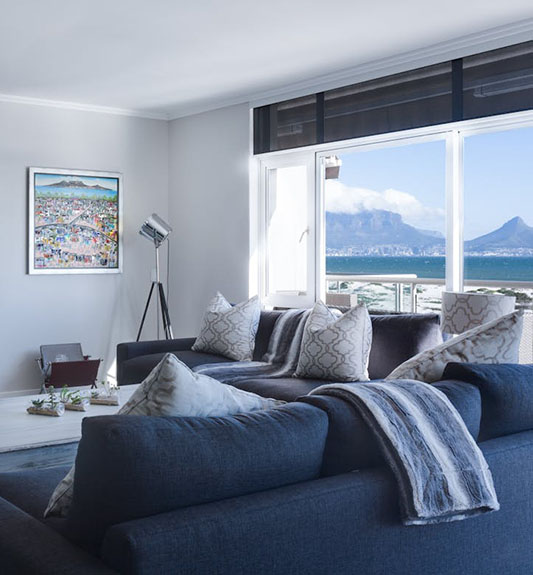 Modern living room featuring a dark sofa with patterned cushions, a scenic ocean view, and a colorful artwork on the wall.