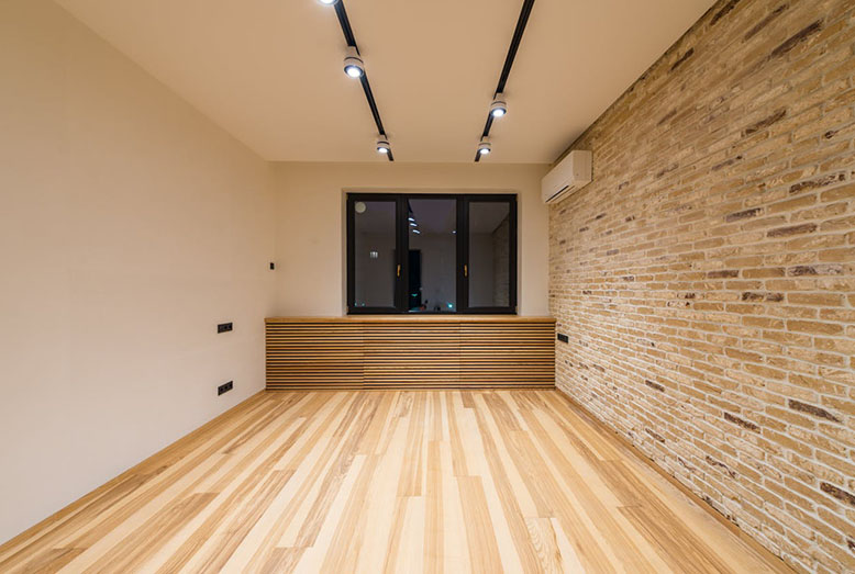 Modern room featuring wooden flooring, exposed brick wall, and a large window with sleek lighting fixtures above.
