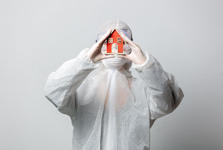 A person in a protective suit holds a small red house model in front of their face, symbolizing safety and care.