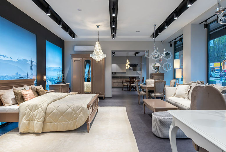 A stylish showroom featuring a modern bedroom with a beige quilt, elegant lighting, chic furniture, and a kitchen area in the background.