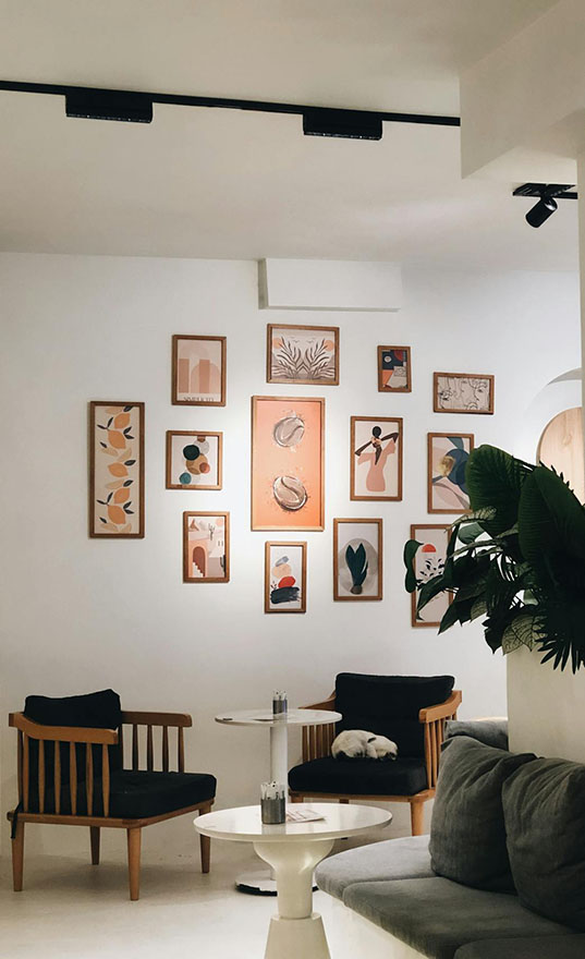 Cozy interior featuring art-filled wall, wooden chairs, a plush couch, and a small table, with a white cat resting nearby.